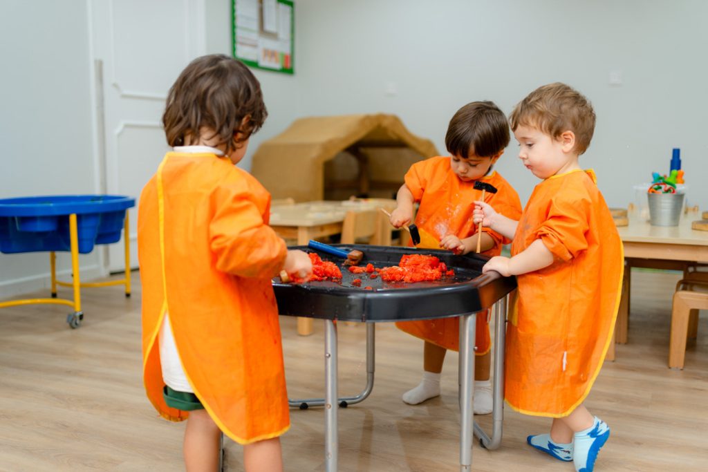 Toddlers wearing aprons using small tools to play with sensory material at a table to improve fine motor skills